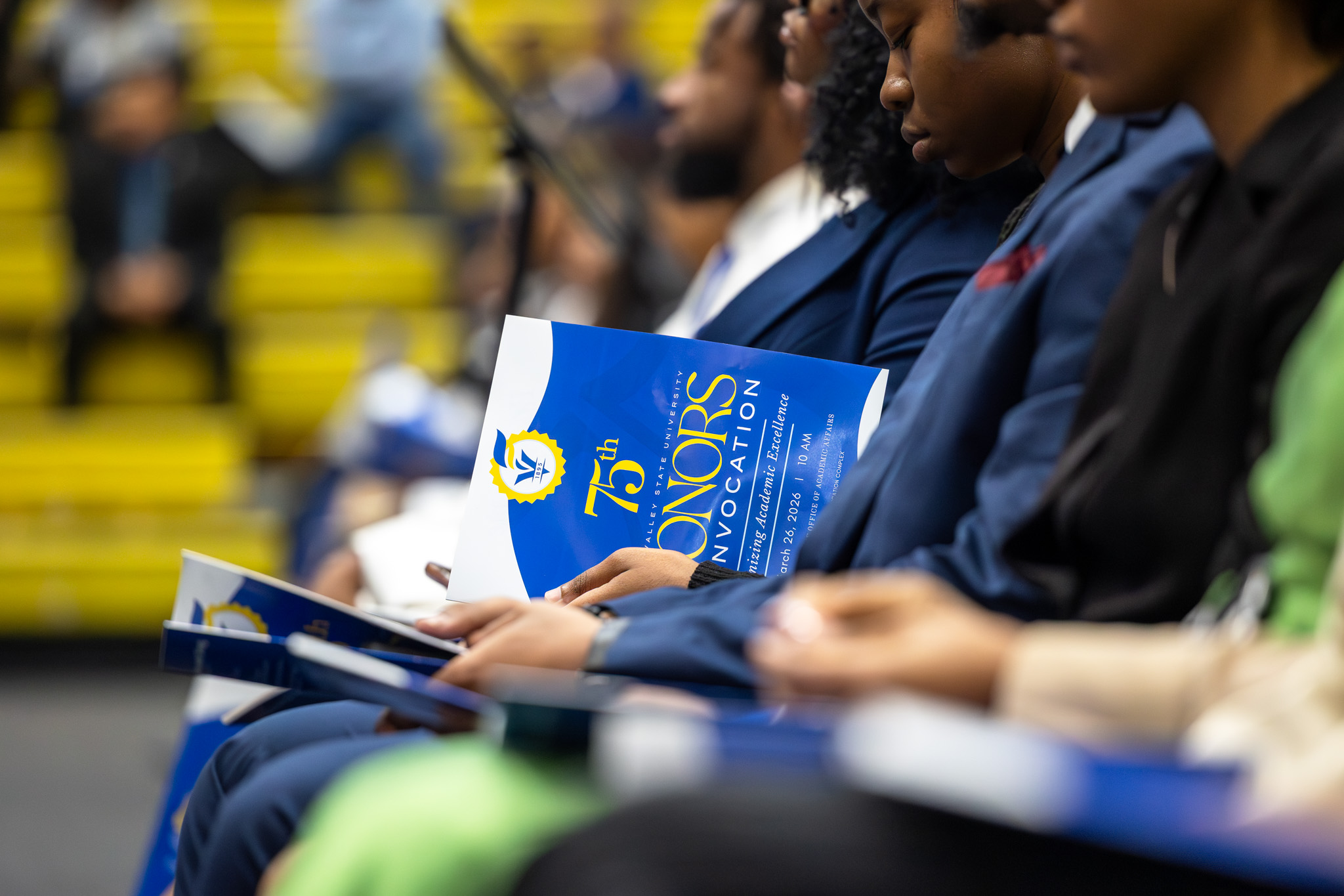 Group of students sitting in chairs in a row with one student looking at the program.