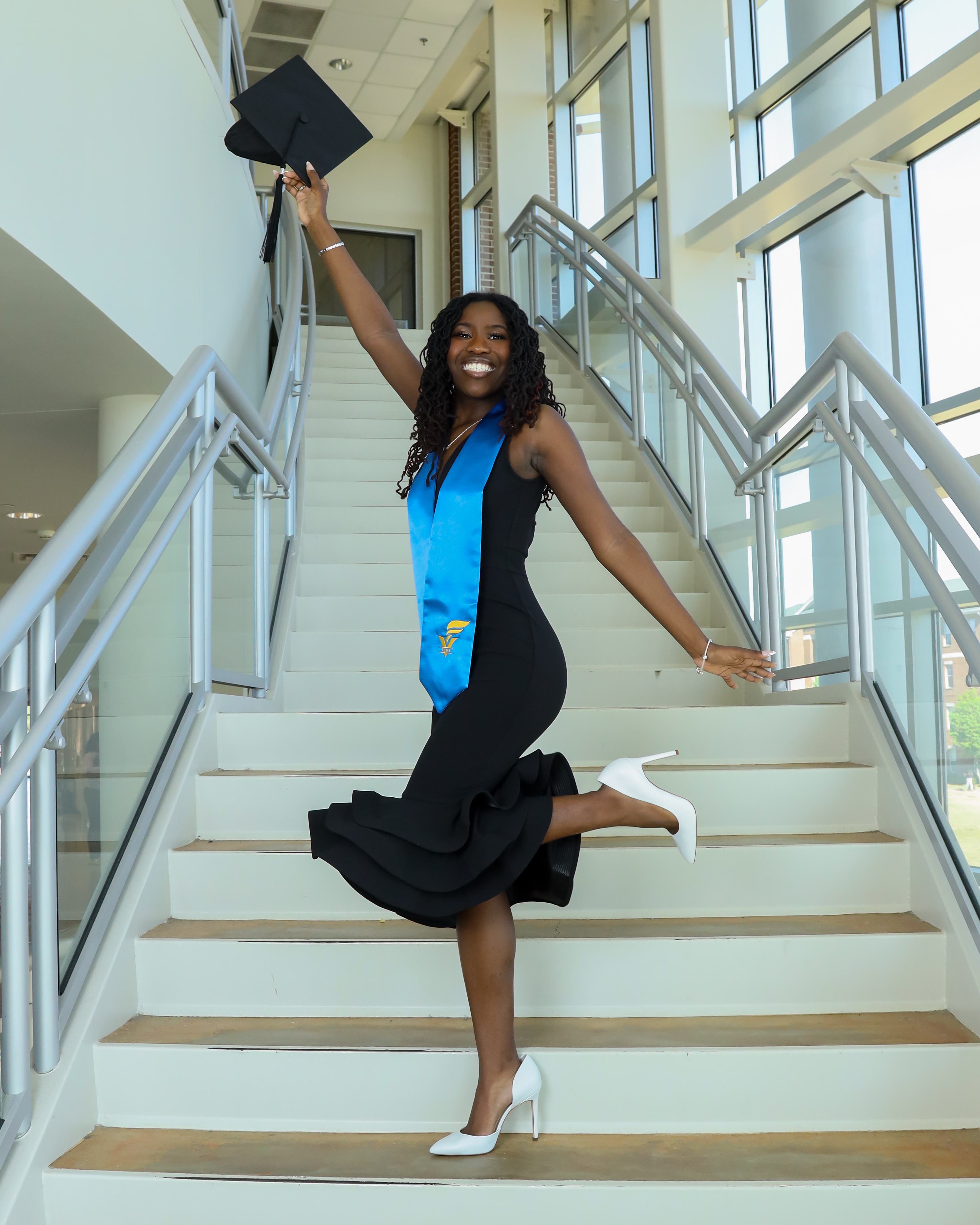 young woman standing on steps with one hand in the air holding a cap and one hand down.